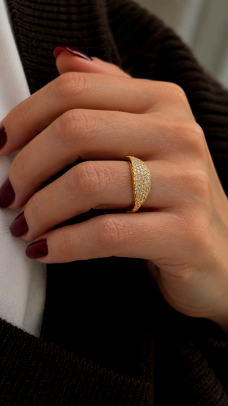 A close-up of a hand wearing a gold ring with pavé-set clear stones, styled with dark burgundy nail polish against a dark brown ribbed sweater and white shirt background.