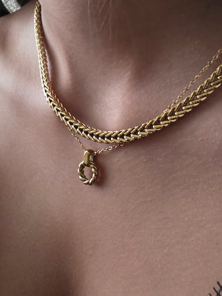 A close-up of a person's neck and upper chest wearing a layered gold necklace. The top layer is a thick braided chain, and the bottom layer is a delicate chain with a small twisted ring pendant.