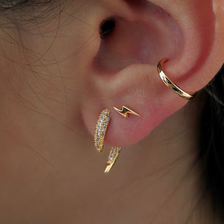 Close-up of a person's ear showcasing a stack of gold earrings, including a gold hoop in the cartilage, a small gold lightning bolt stud, and two gold huggie hoop earrings set with clear pavé stones.