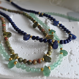 A close-up of several beaded bracelets arranged on a white textured display stand. The bracelets feature dark blue beads, light green translucent beads, and earthy brown/green marbled beads with small gold spacer accents.