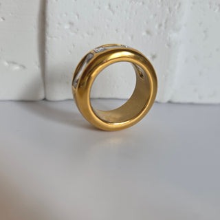 A gold ring with a textured, organic band shape resting on a white textured surface.