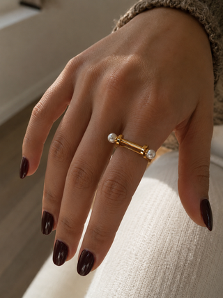 A close-up of a hand wearing a gold ring with two small white pearl-like beads, set against a soft neutral indoor background.