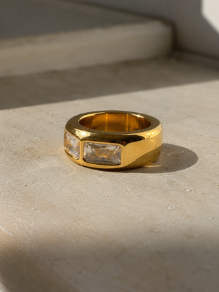 A wide gold ring with three rectangular clear gemstones baguette cut Cubic zirconia, resting on a textured beige stone surface in natural light.