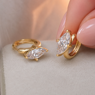 A pair of gold hoop earrings with marquise-cut clear moissanite gemstones, with one ring being held by a hand with pink manicured nails against a soft beige background.