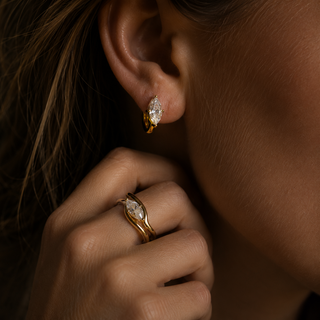 A close-up lifestyle shot of a model wearing a gold hoop earring with a marquise-cut clear moissanite gemstone and a matching gold moissanite ring on her finger.