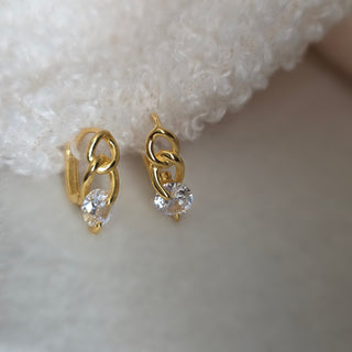 A pair of small gold huggie hoop earrings with a clear heart-shaped crystal drop, resting on a soft white textured fabric.