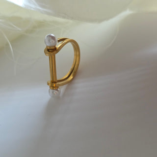 A gold ring featuring a vertical bar with two small white pearl-like beads, one on each end, set against a soft white background with delicate white feathers.