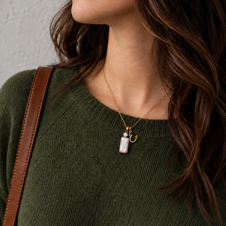 A close-up lifestyle shot of a woman wearing a gold necklace with a white rectangular pendant and a small gold script charm, paired with an olive green knit sweater and a brown leather shoulder strap.