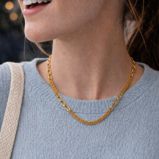 A close-up lifestyle shot of a woman wearing a delicate gold chain necklace with oval links, paired with a light blue knit sweater.
