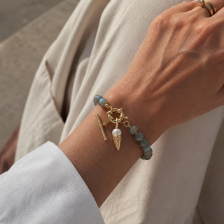 A close-up of a person's wrist wearing a delicate aventurine beaded charm holder bracelet. The bracelet features light blue-green beads, a gold toggle clasp, a small gold ring, a white pearl icecream charm, and a gold Tbar charm attached to the clasp