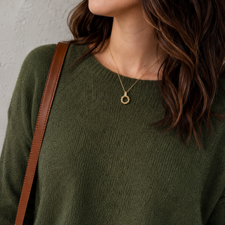 A woman wearing an olive green knit crewneck sweater, blue jeans, and a gold necklace, with a brown shoulder bag strap visible.