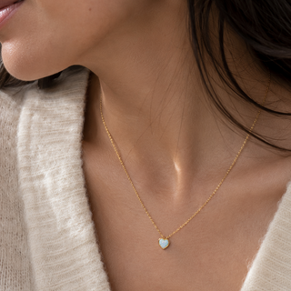 A close-up of a model wearing a delicate gold chain necklace with a small Opal gemstone heart-shaped pendant that has a white iridescent center, styled with a cream-colored knit top.