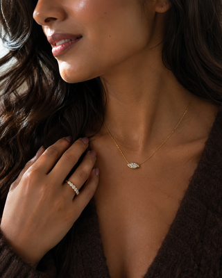 A close-up lifestyle shot of a woman wearing a delicate gold necklace with a small white crystal-encrusted pendant, paired with a matching crystal ring on her finger.