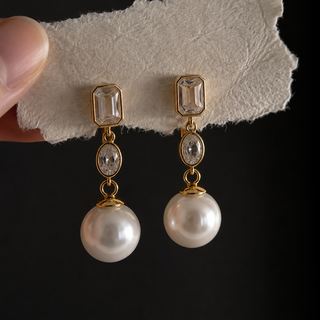 A pair of gold drop earrings with clear sparkly rectangular and oval moissanite gemstones, each suspending a large ivory white round pearl, held against a black background on a textured white card with a finger visible on the left.