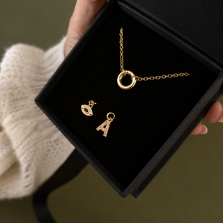 A gold jewelry set including a charm clasp holder necklace, charms, and letter pendant, presented in an open black gift box held by a hand wearing a cream knit sleeve.